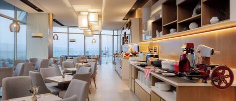 A modern dining area with tables and chairs, a kitchen counter with appliances, and shelves with decorative items.