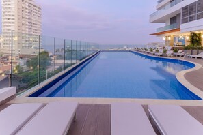 Une piscine à débordement située sur le toit, avec des chaises longues, une balustrade en verre et une vue sur le paysage urbain.