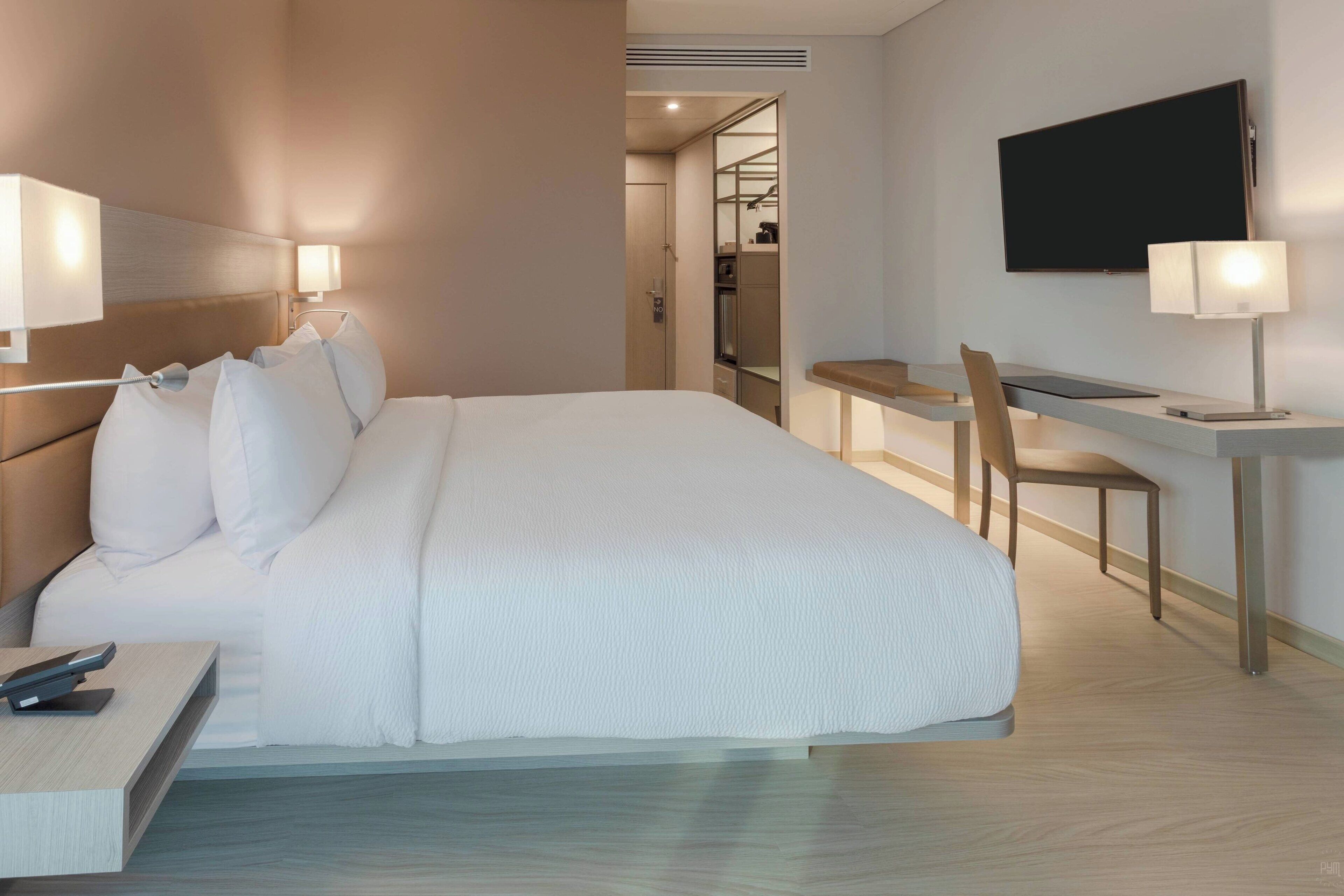Ein Hotelzimmer mit einem großen Bett, einem Schreibtisch, einem Sessel, einem an der Wand befestigten Fernseher und einem begehbaren Kleiderschrank.