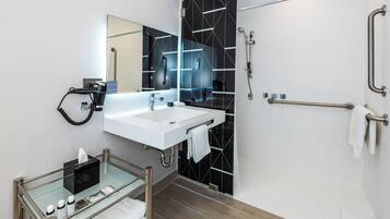 A modern bathroom with a glass-topped vanity, a white sink, a large mirror, and a black tiled shower area.