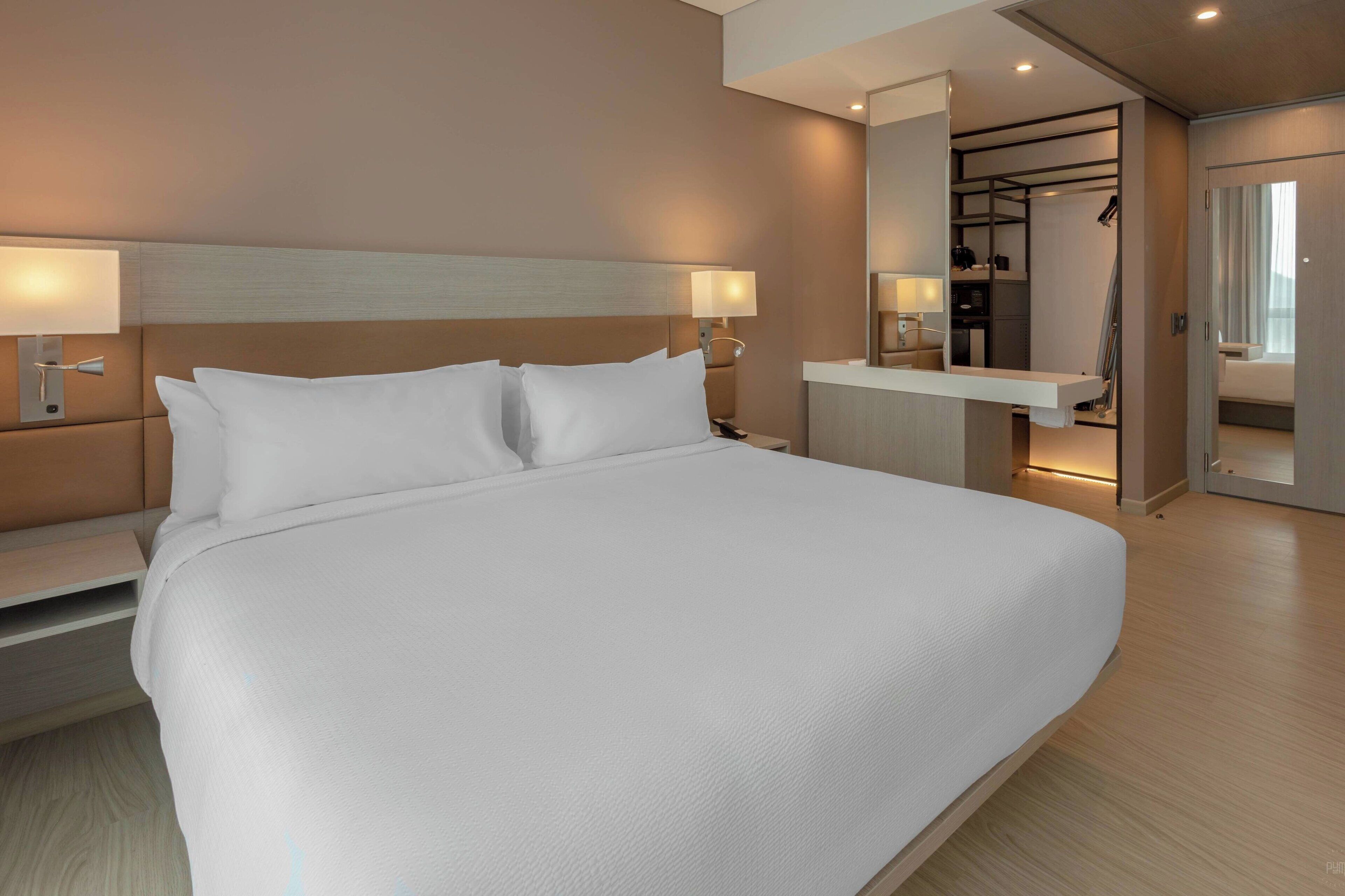 A modern hotel room with a large bed, bedside tables, a built-in desk, and a mirror.