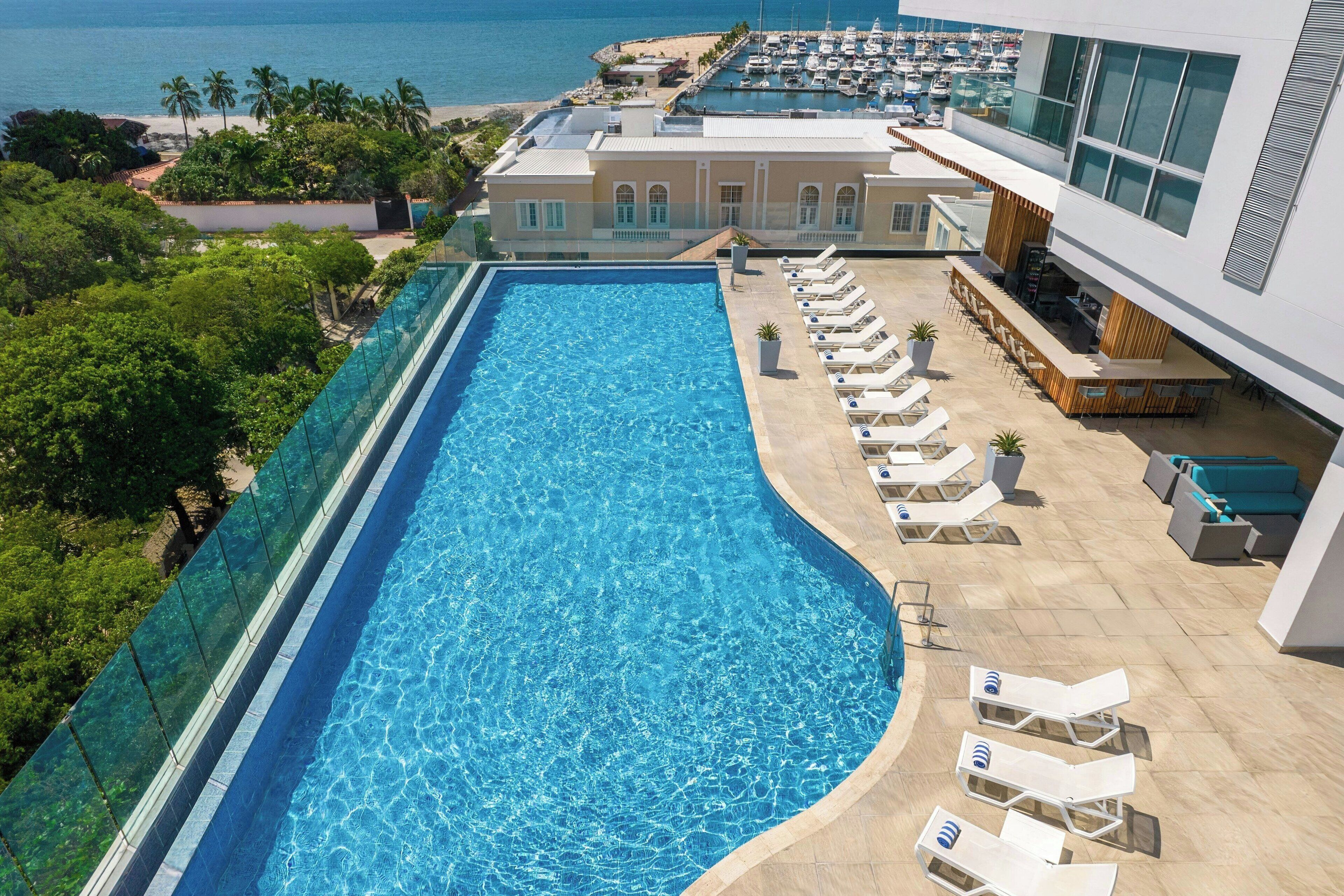 A rooftop pool with clear blue water, surrounded by lounge chairs and a view of the ocean and marina.