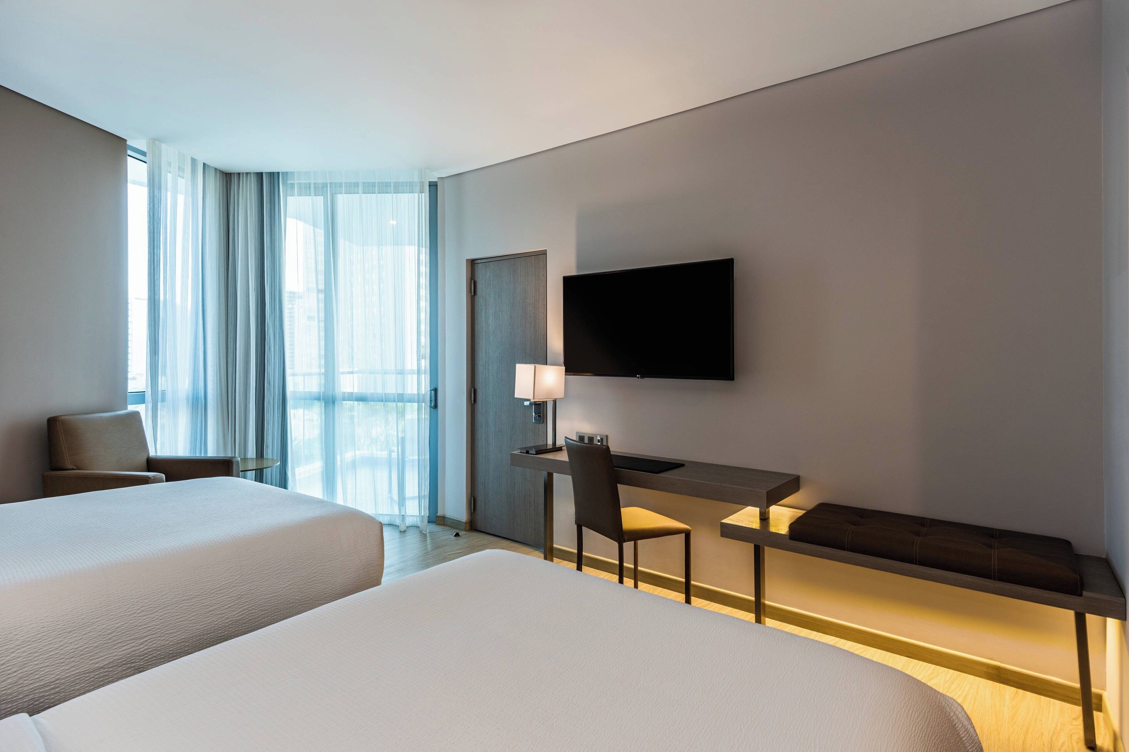 A hotel room with two beds, a desk with a chair, a TV mounted on the wall, and a large window with curtains.
