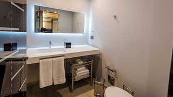 Un baño moderno con lavamanos, espejo y retrete blancos. Hay toallas colgadas en un perchero y un cubo de basura.