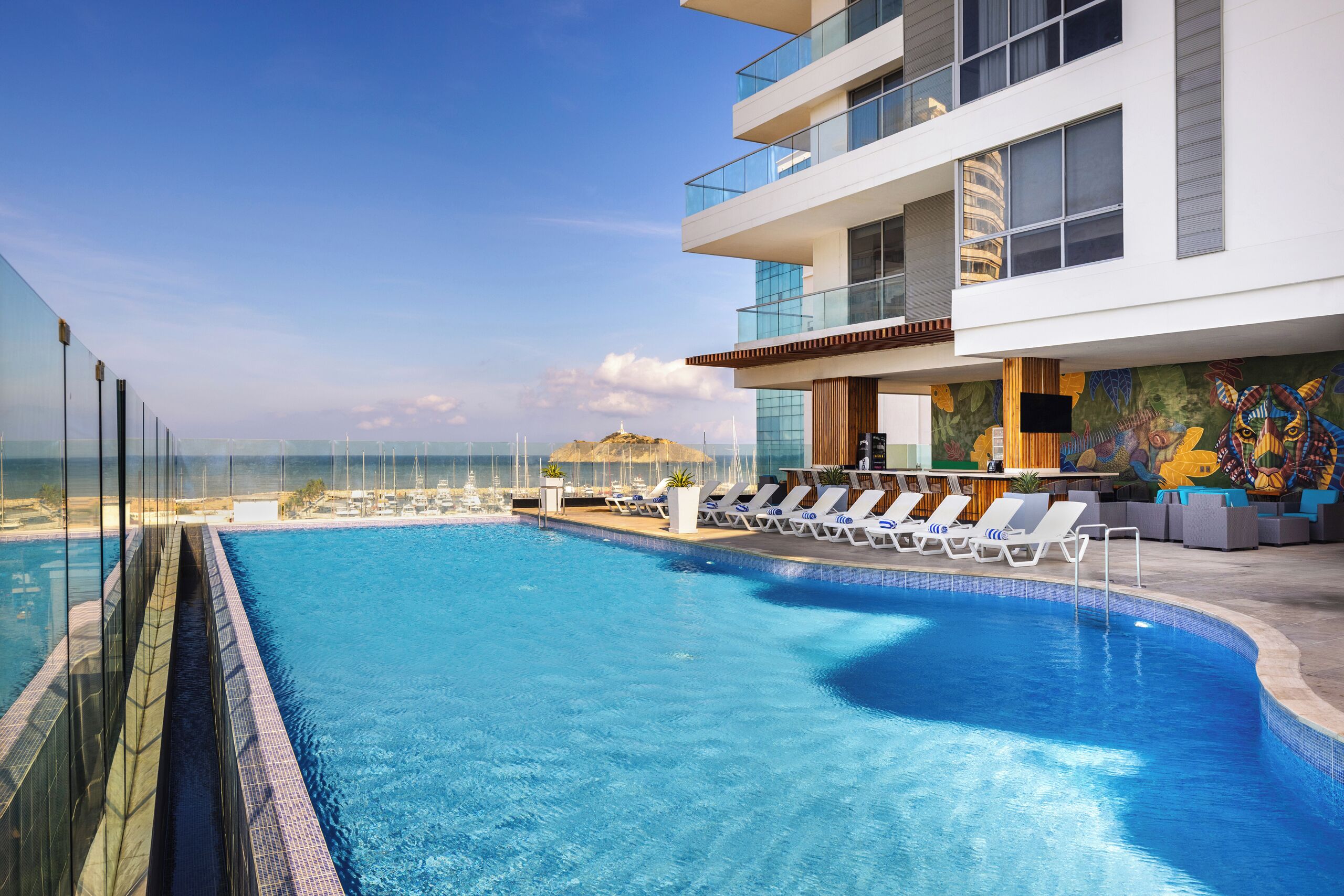 A modern hotel with a rooftop infinity pool overlooking the ocean, lounge chairs, and a colorful mural.