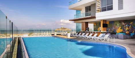 A modern hotel with a rooftop infinity pool overlooking the ocean, lounge chairs, and a colorful mural.