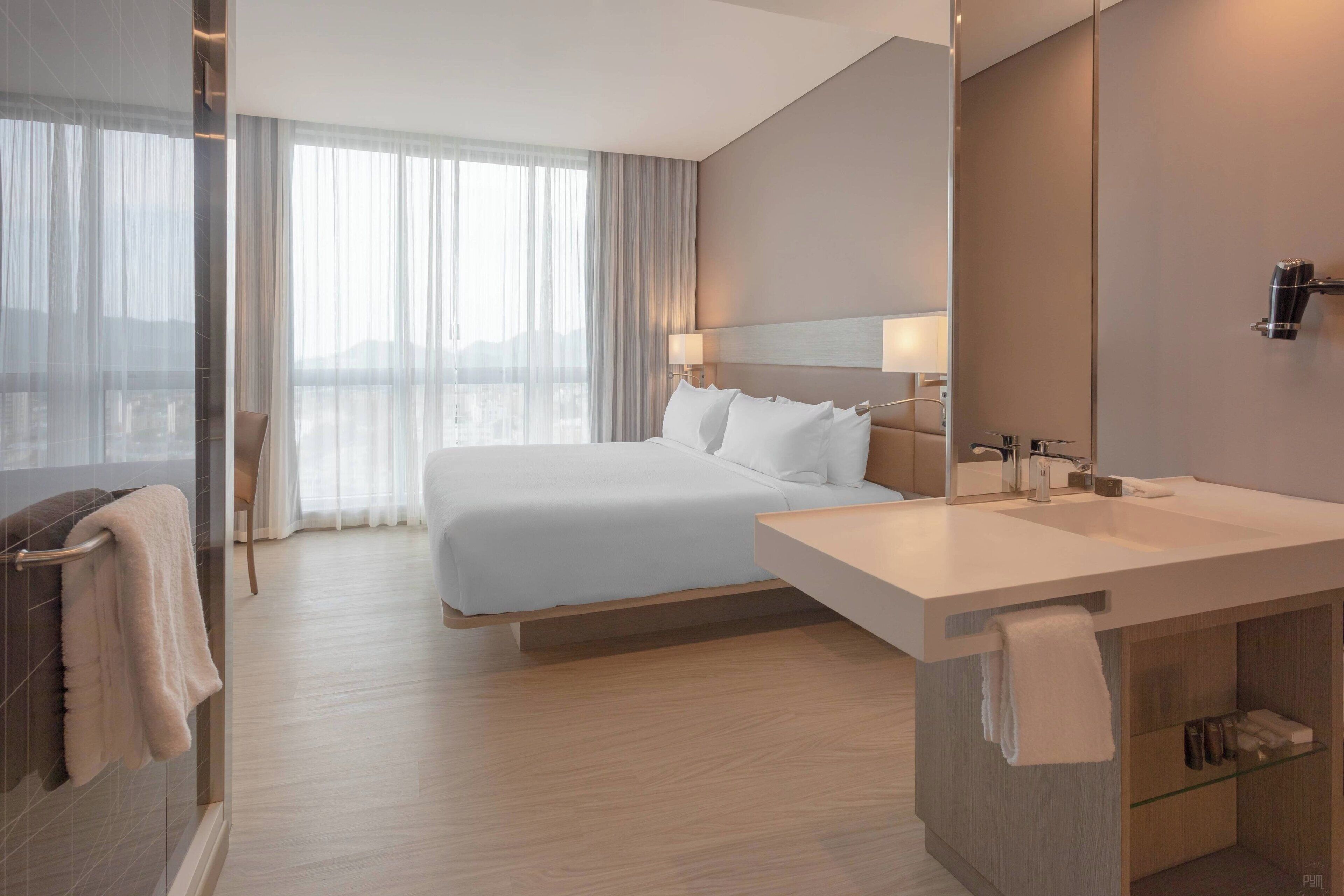A modern hotel room with a large bed, a bathroom with a vanity, and a large window with sheer curtains.