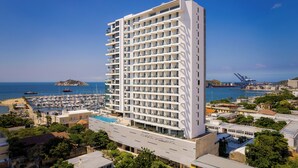 A tall, modern hotel with a white facade, located near a marina with boats and a distant island. - AC Hotel by Marriott Santa Marta (Santa Marta)