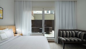 A hotel room with a bed, a sofa, a nightstand, and a view through a glass door to an outdoor area with furniture.