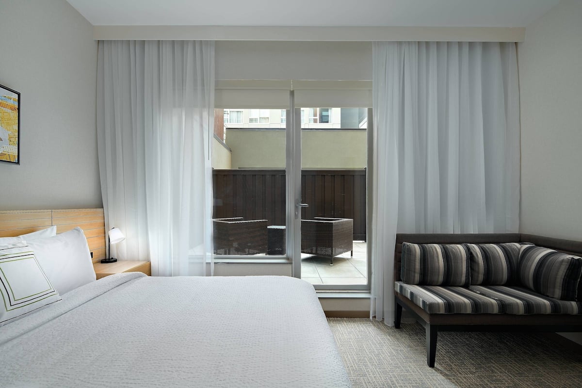 A hotel room with a bed, a sofa, a nightstand, and a view through a glass door to an outdoor area with furniture.