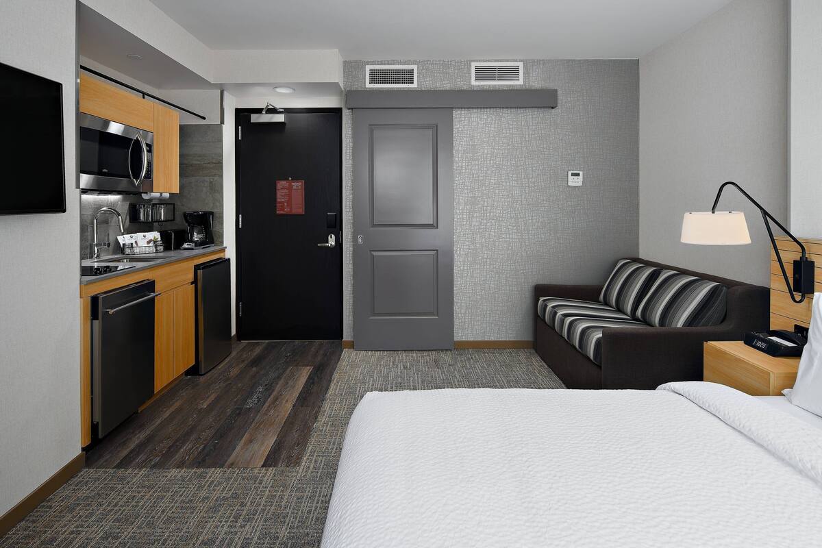 A compact hotel room with a kitchenette, a bed, a sofa, and a bedside table with a lamp.