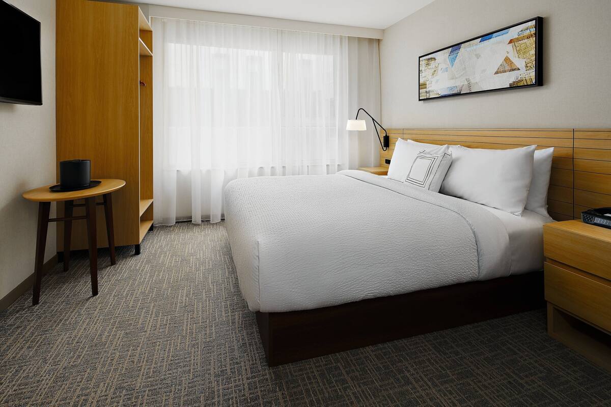 A hotel room with a large bed, a bedside table, a chair, a desk, a lamp, and a painting on the wall.