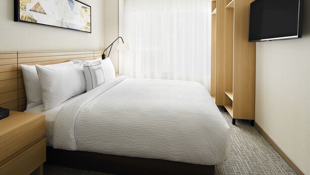 Una habitación de hotel con una cama grande, un televisor de pantalla plana, una mesita de noche, una lámpara y una ventana con cortinas translúcidas.