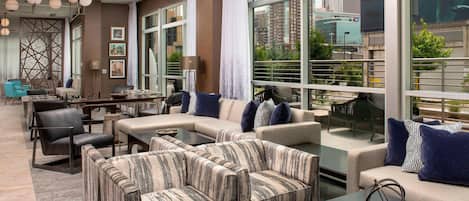 A modern hotel lobby with a sectional sofa, armchairs, a coffee table, and a dining area. Large windows offer views of the cityscape.