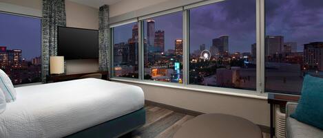 A modern hotel room with a large bed, a seating area, and a city view through large windows.