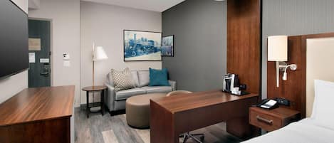 Ein Hotelzimmer mit einem Holztisch, einer Couch mit Kissen, einem Bett, einem Nachttisch mit Lampe und einem gerahmten Stadtbild an der Wand.
