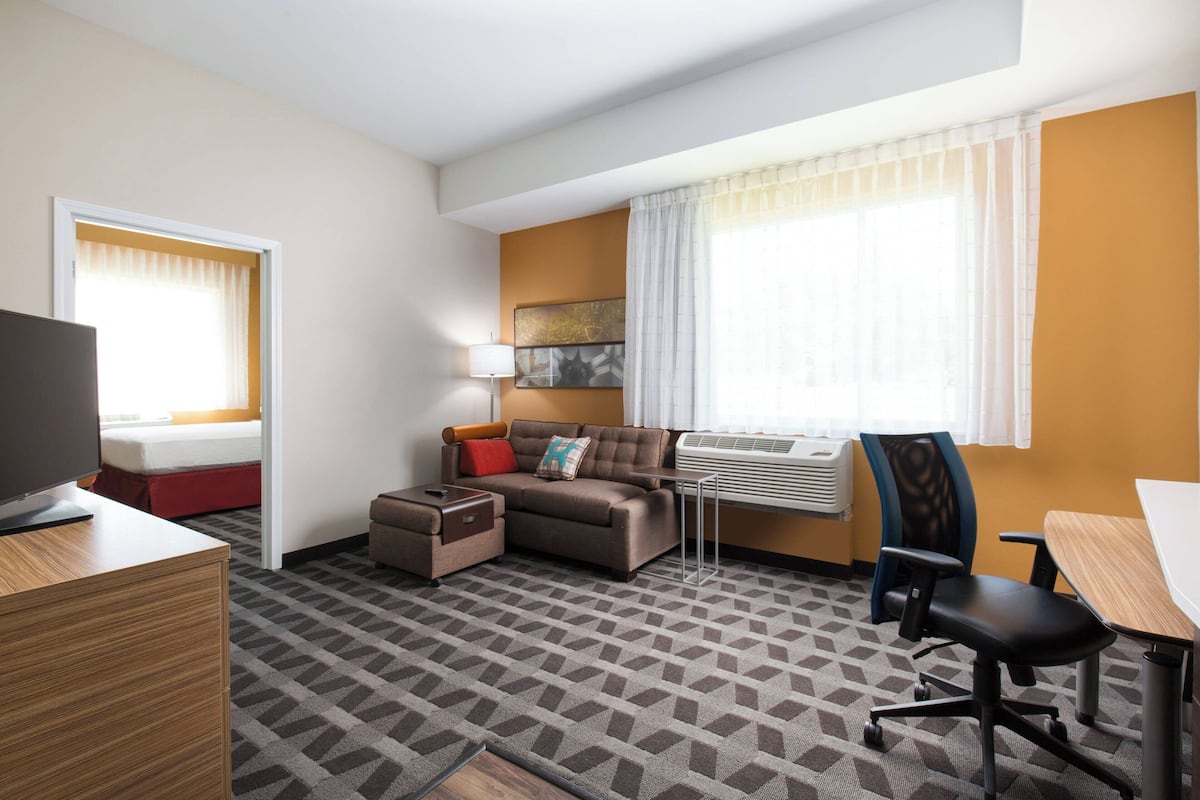 Habitación de hotel con sofá, escritorio, silla, televisor y ventana con cortinas.