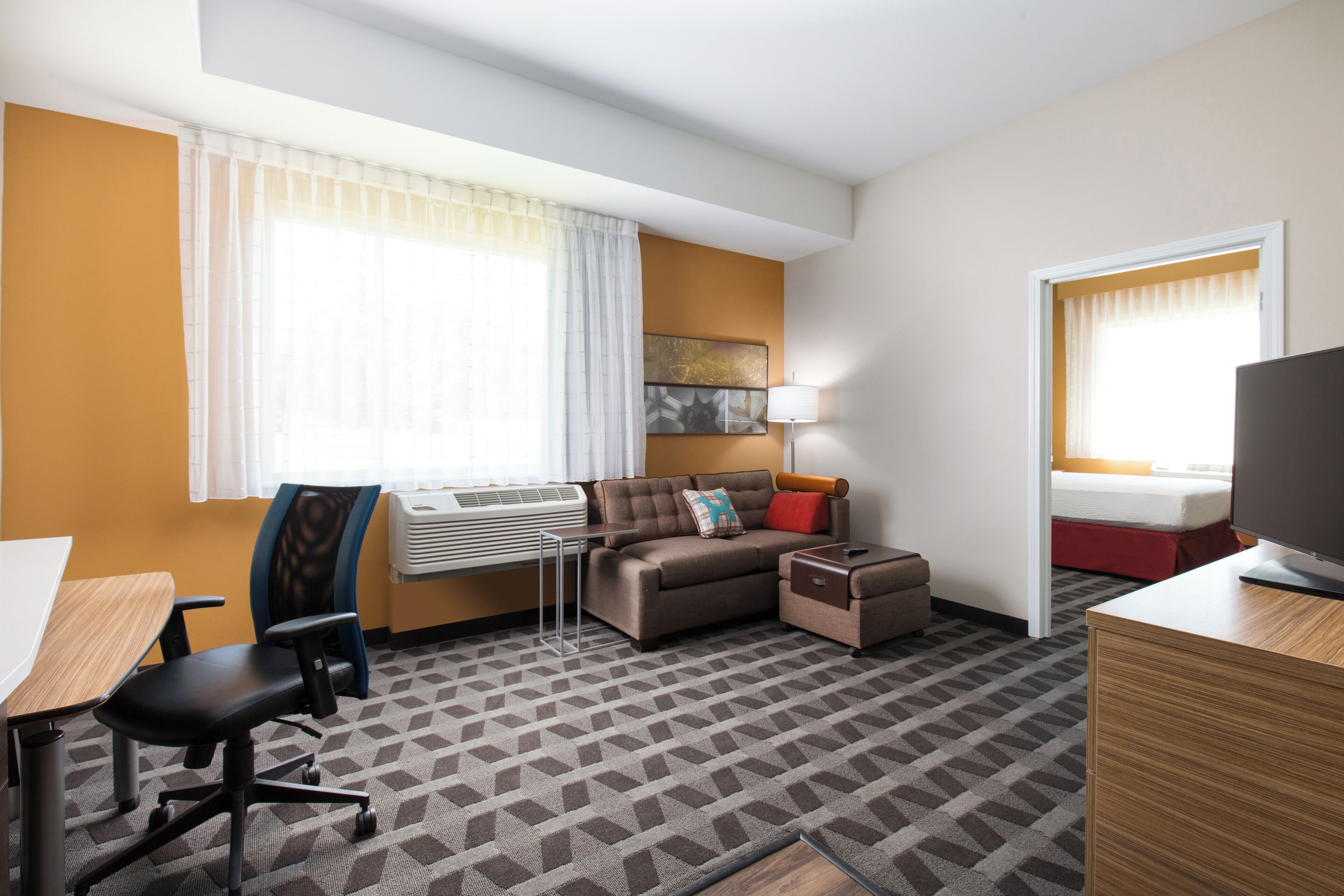A hotel room with a sofa, a desk, a chair, a TV, and a window with curtains.