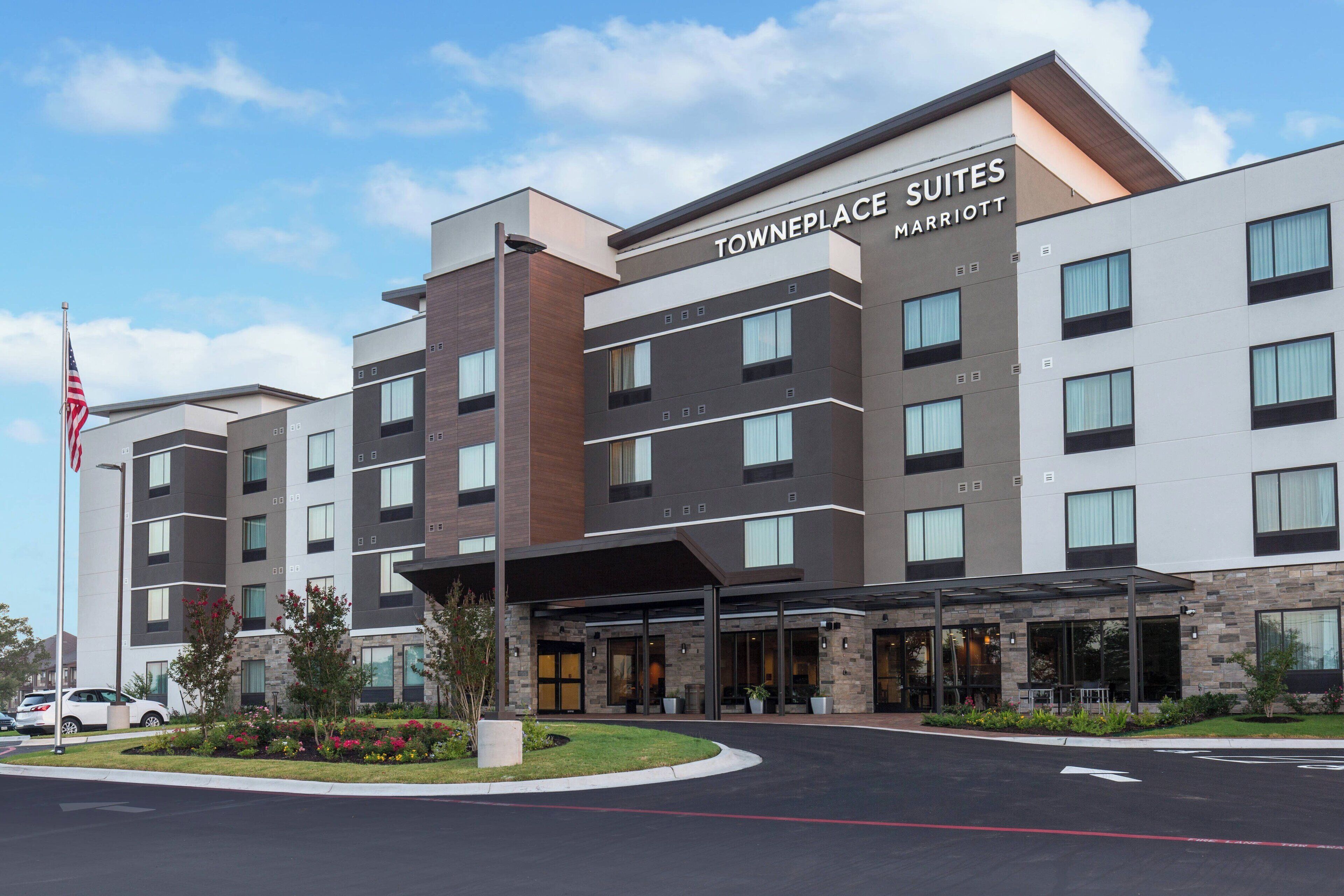 Egy modern hotel, amelyen jól láthatóan szerepel a „Towneplace Suites Marriott” felirat. Többszintes épület, üveg- és téglaelemekkel kombinált homlokzattal, fedett bejárattal, valamint gondozott előtérrel, növényekkel és zászlórúddal.