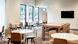Uma sala de jantar de hotel com mesas redondas, cadeiras e uma grande tela de televisão na parede.