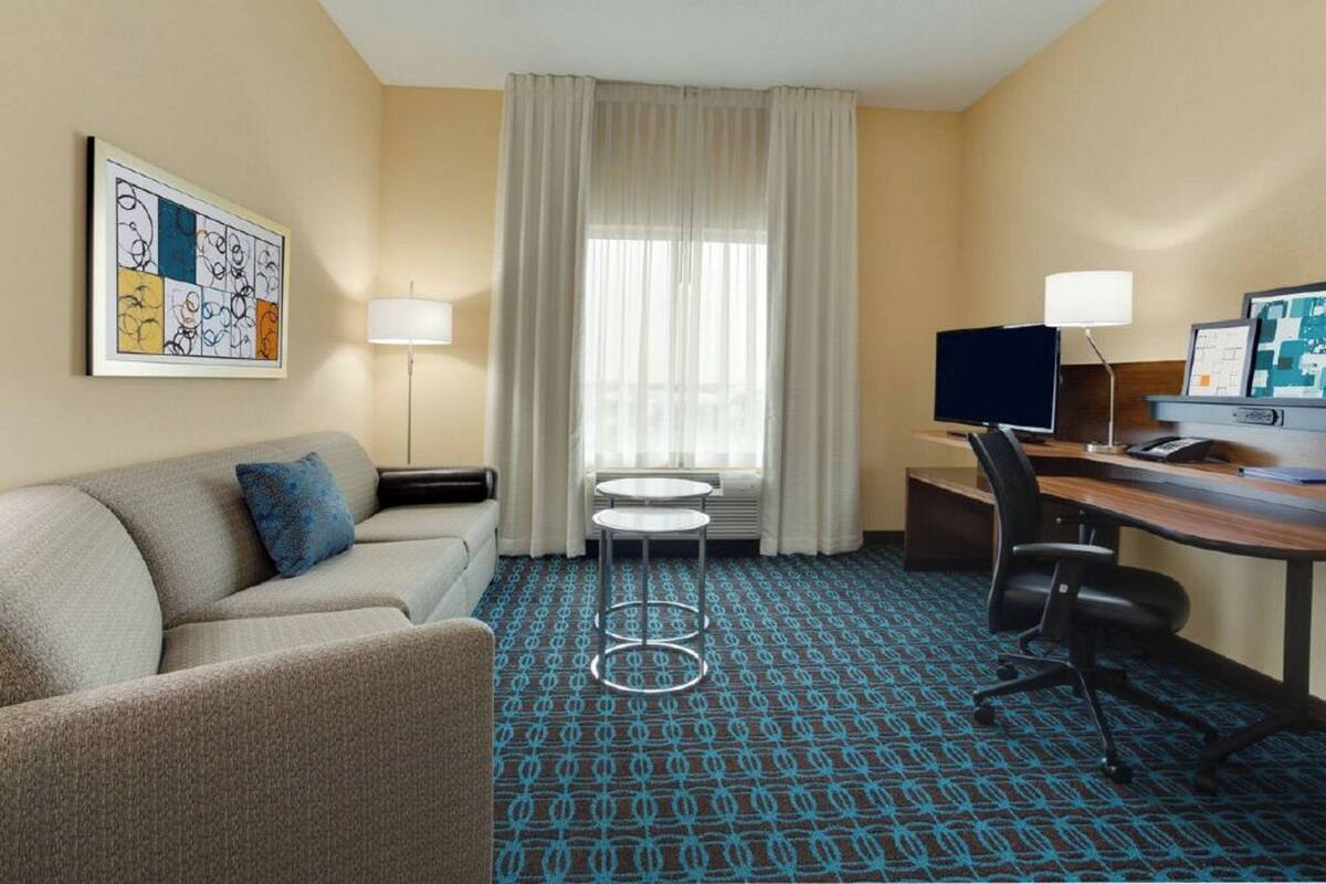 Une chambre d’hôtel avec un canapé, un bureau avec un ordinateur, une petite table et une fenêtre avec des rideaux.