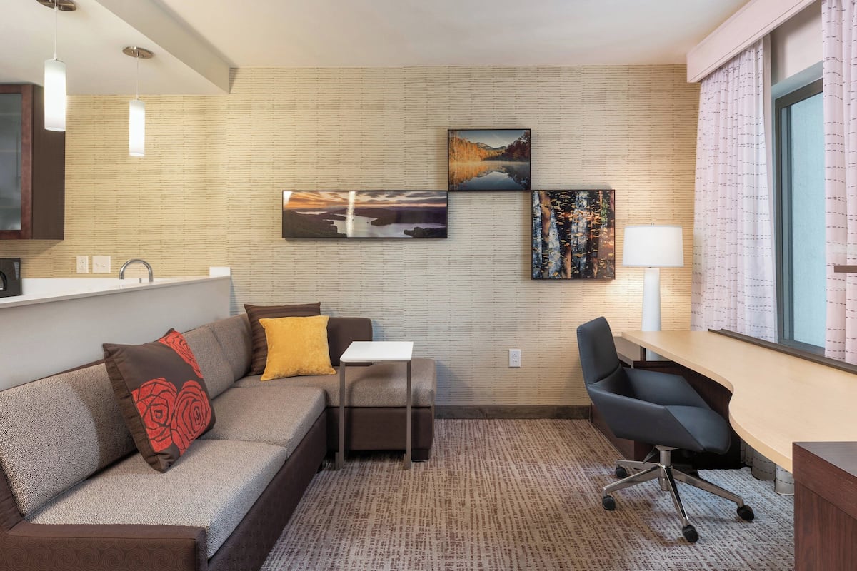 A modern hotel room with a sofa, a small desk, a chair, and artwork on the wall.