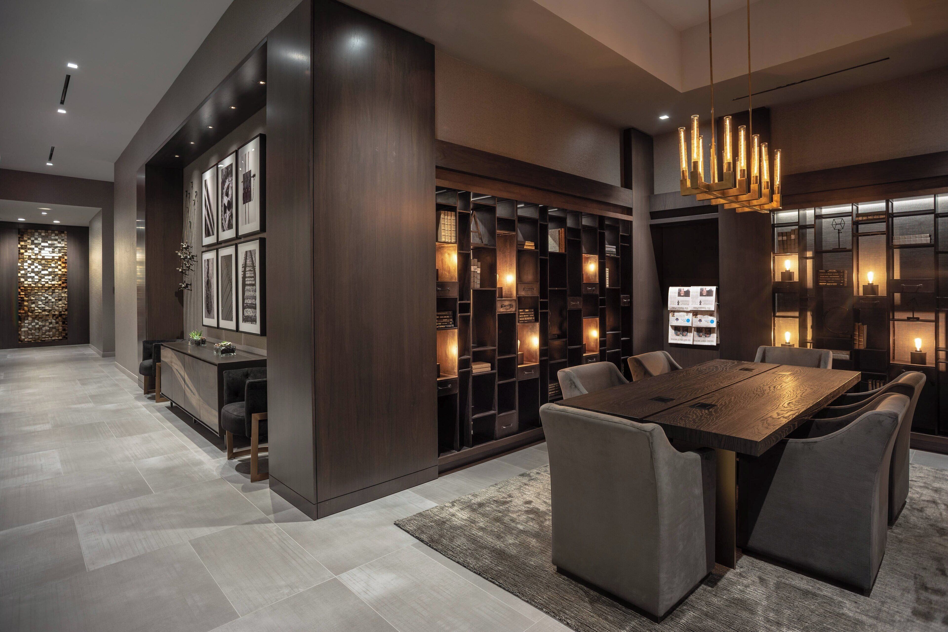 A modern dining area with a large wooden table and upholstered chairs, surrounded by dark wood shelving with illuminated sections.