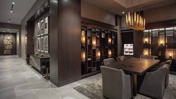 A modern dining area with a large wooden table and upholstered chairs, surrounded by dark wood shelving with illuminated sections.