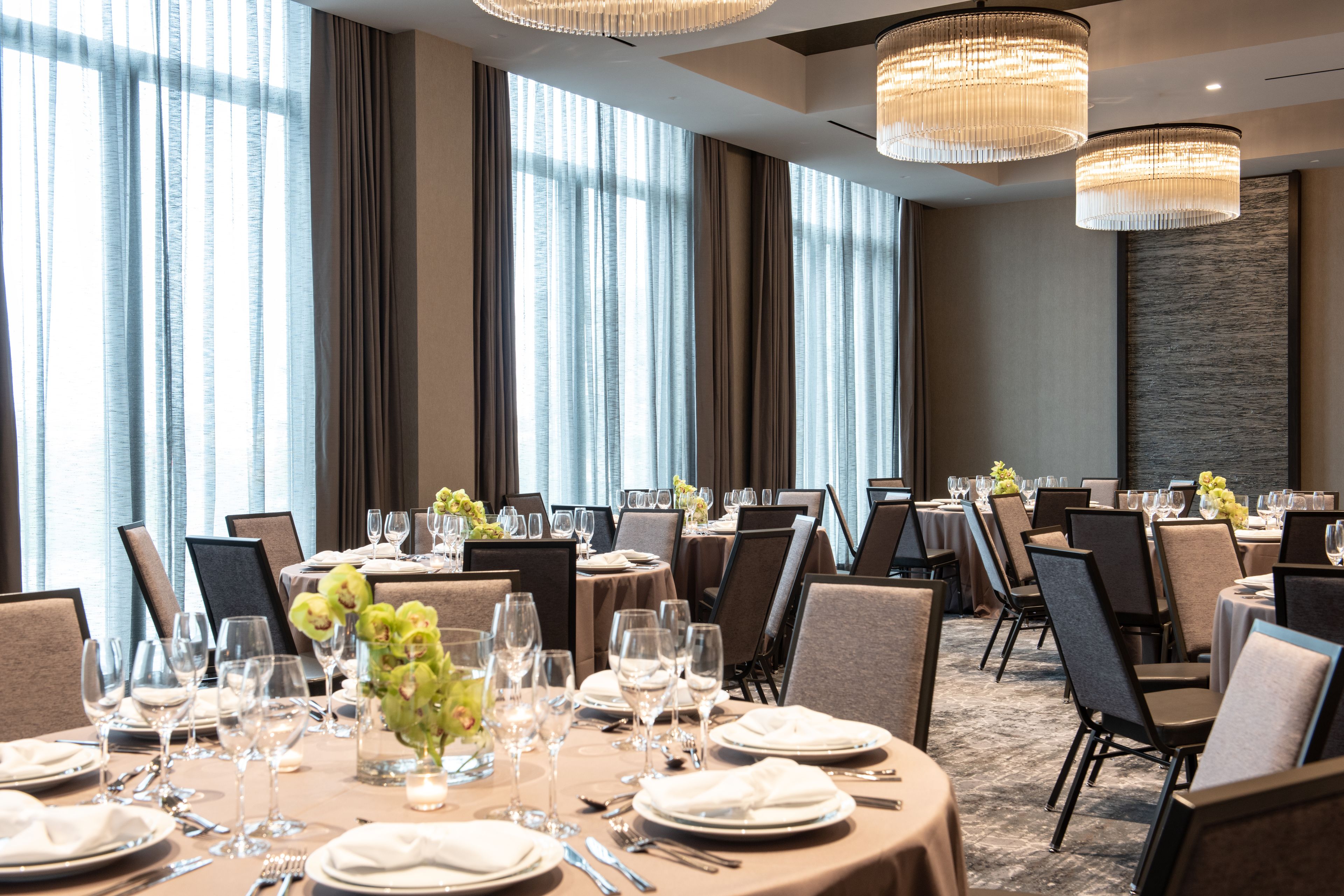 A dining area with round tables set for a meal, large windows with curtains, and modern chandeliers.