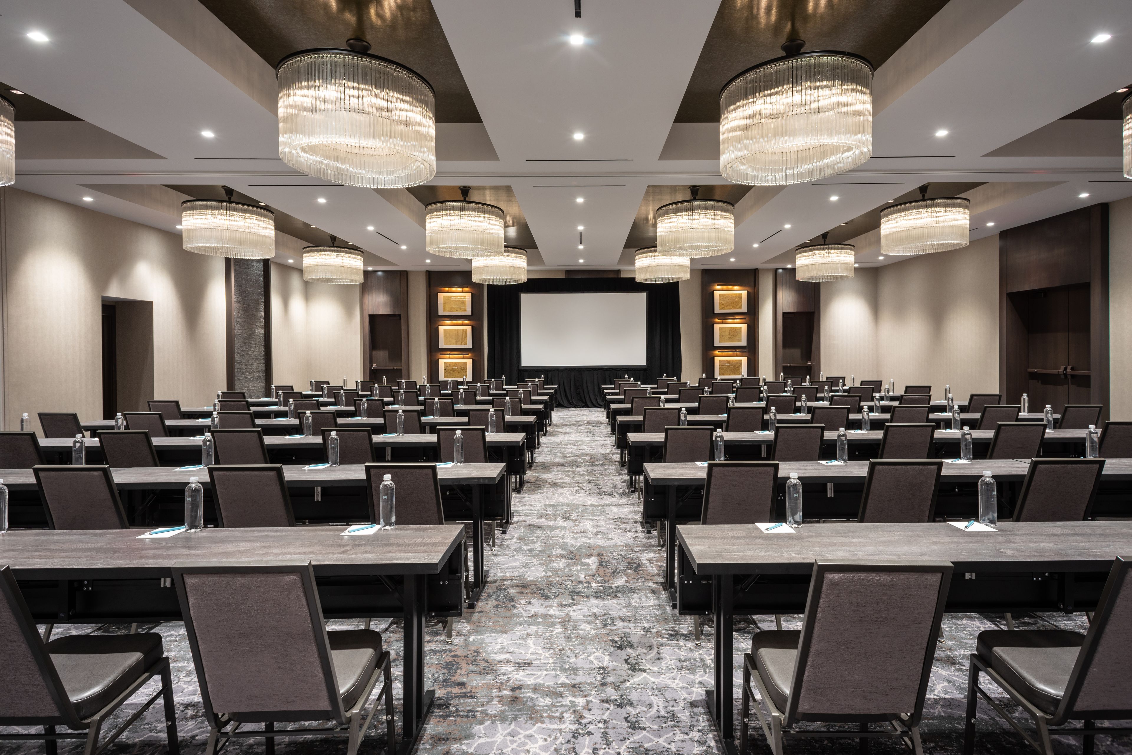 A conference room with rows of tables and chairs, large chandeliers, and a projector screen.