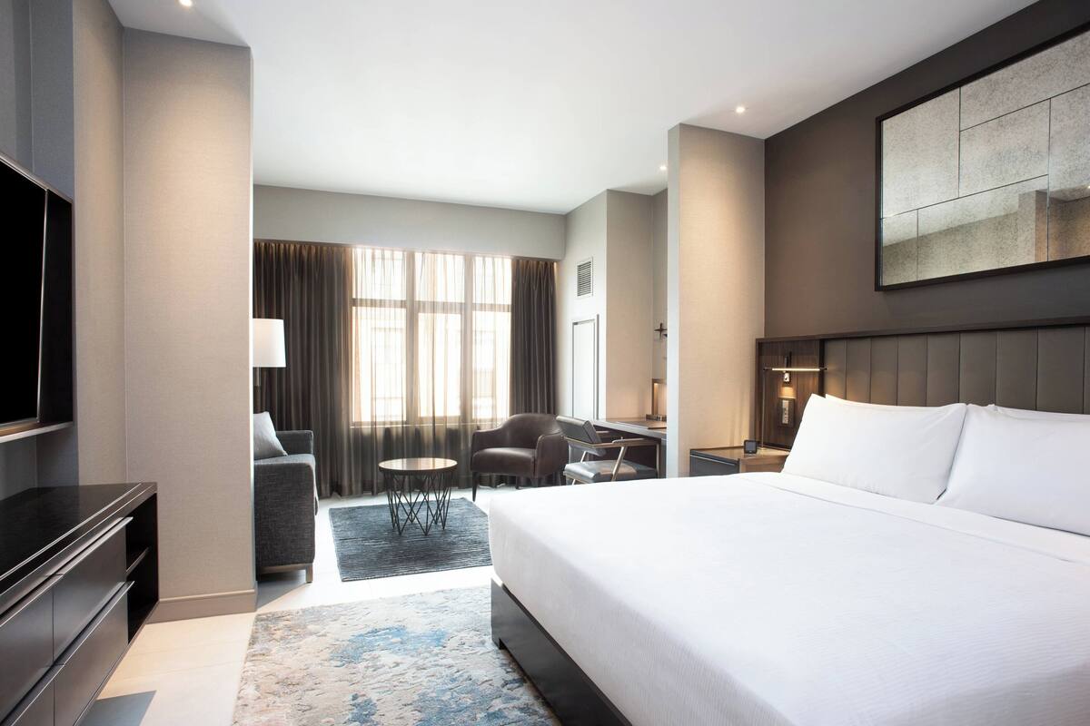 Ein modernes Hotelzimmer mit einem großen Bett, einem Schreibtisch, einem Sessel, einem Fernseher und einem großen Fenster mit Vorhängen.