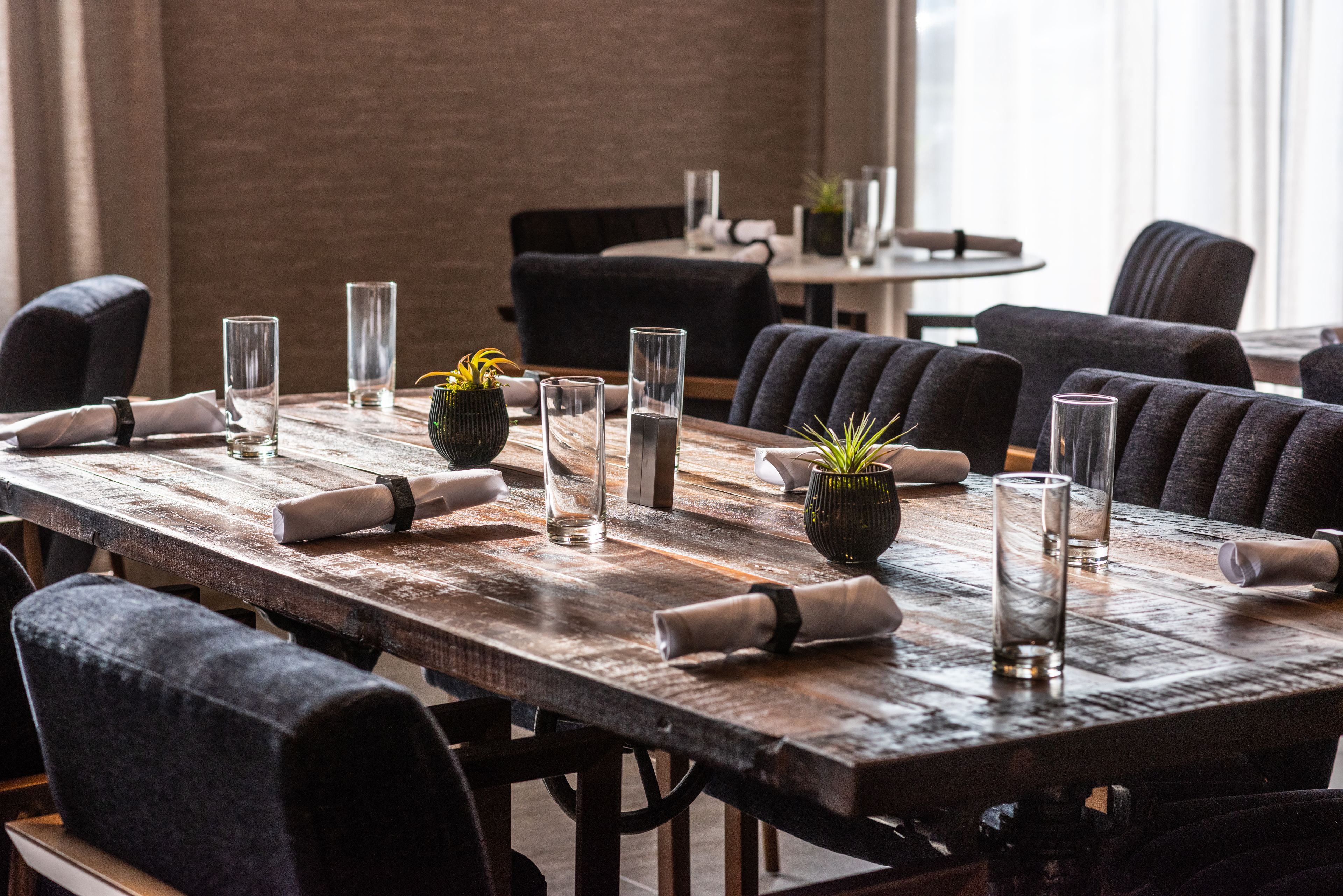 A long wooden dining table set with glassware, napkins, and small potted plants, surrounded by upholstered chairs in a modern dining area.