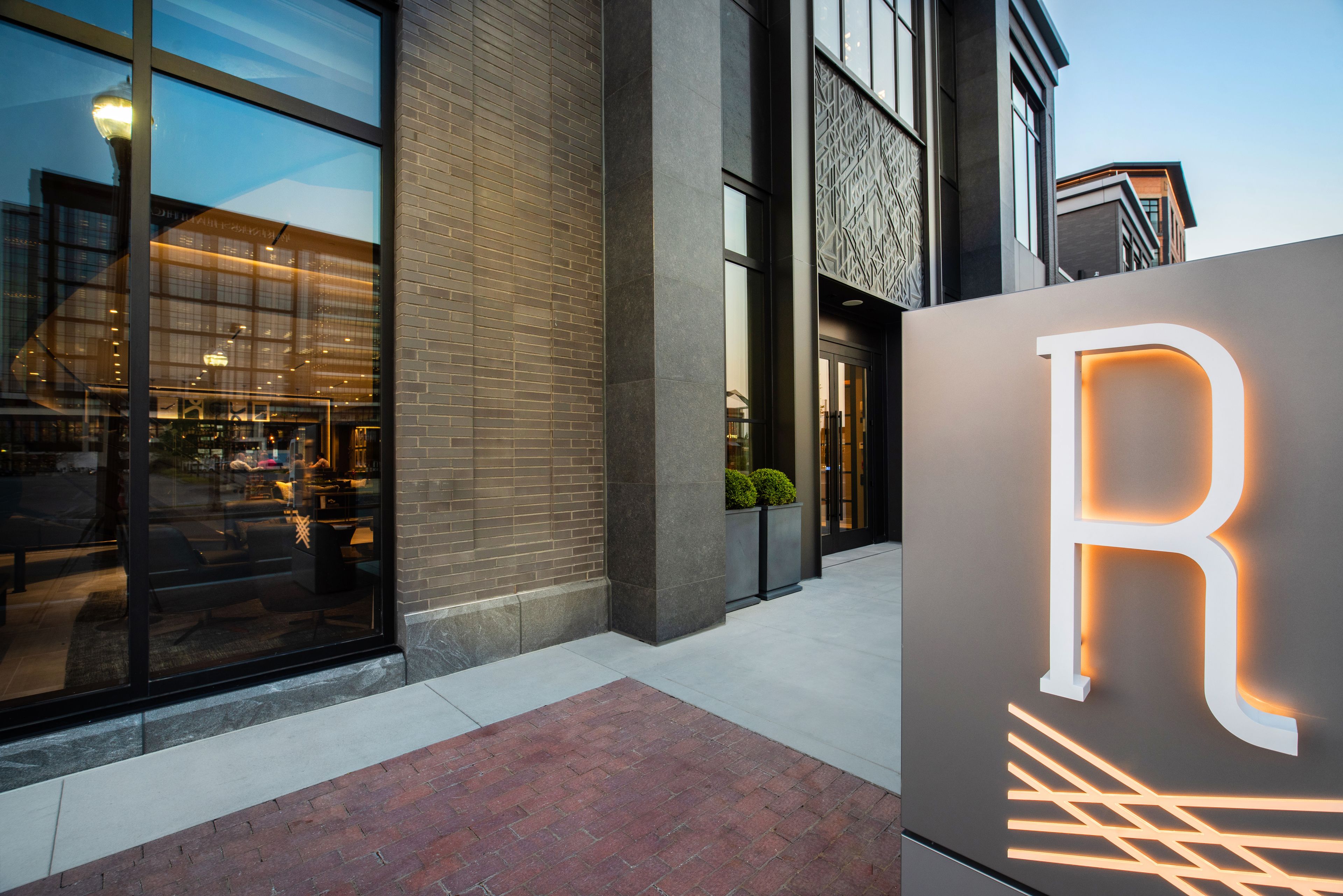 A modern building with a prominent illuminated 'R' sign, large windows, and a brick-paved entrance.