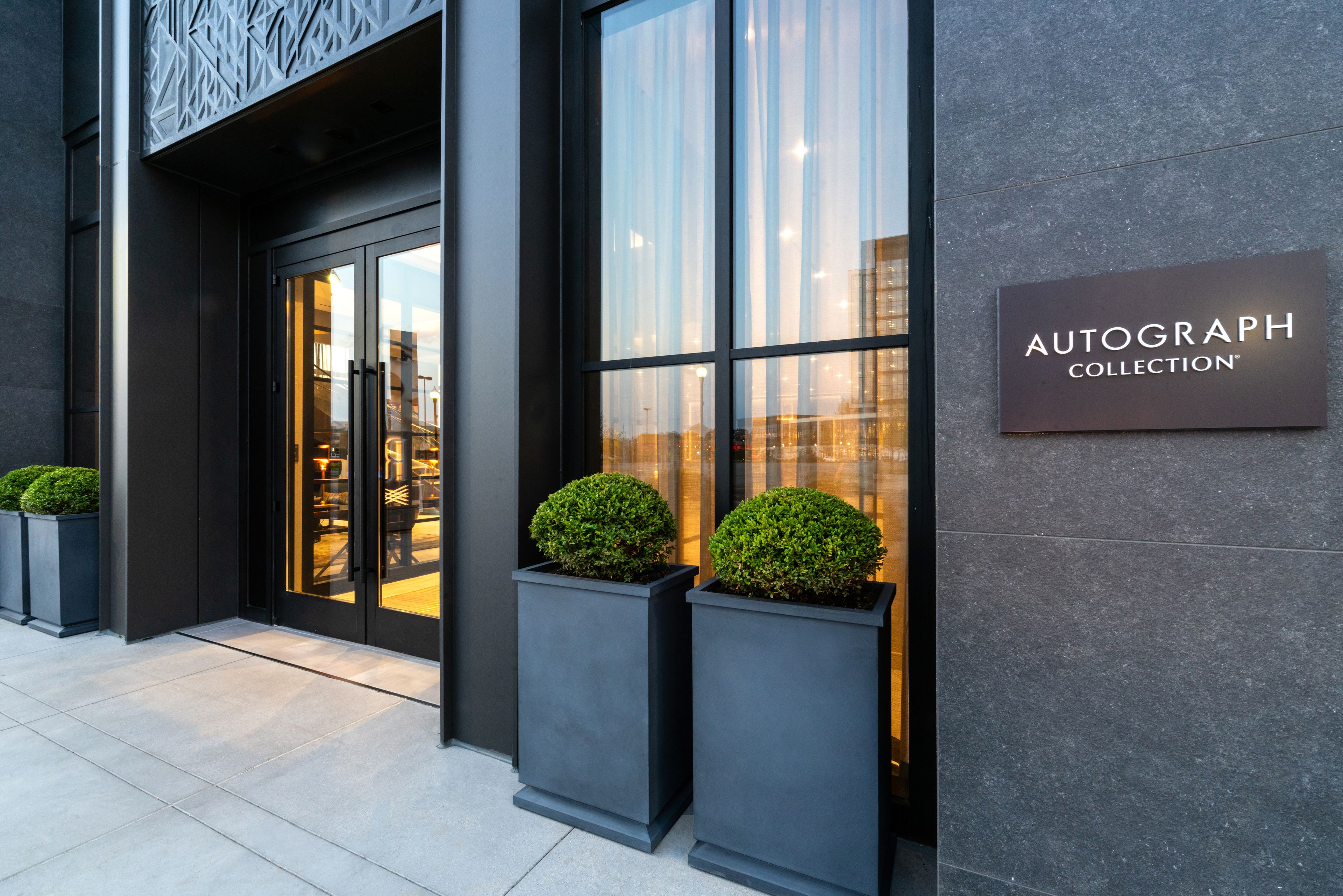 A modern building entrance with a glass door, two potted plants, and a sign reading "AUTOGRAPH COLLECTION".