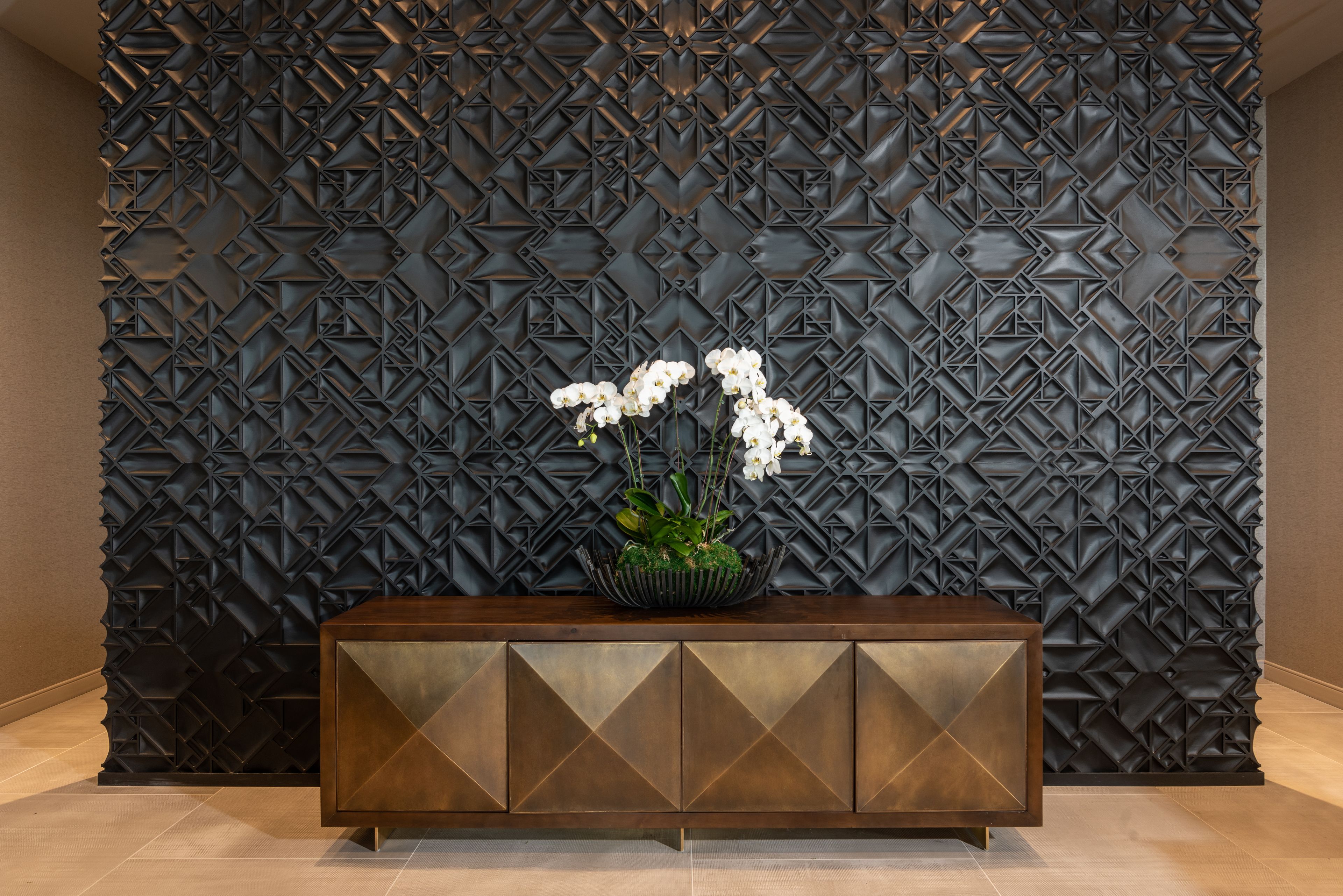 Un espace d’accueil moderne avec un mur orné d’un motif géométrique, une console en bois et une plante en pot aux fleurs blanches.