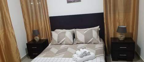 A neatly made bed with a patterned bedspread, two pillows, and a rolled towel on the bed. There are two nightstands with lamps and a small picture on the wall above the bed.
