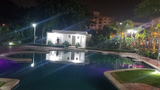 Vue nocturne d’un espace piscine avec un petit bâtiment, entouré de palmiers et bien éclairé par des lampadaires.