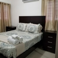 Una cama cuidadosamente tendida con ropa de cama estampada, un cabecero oscuro y dos mesitas de noche con cajones.