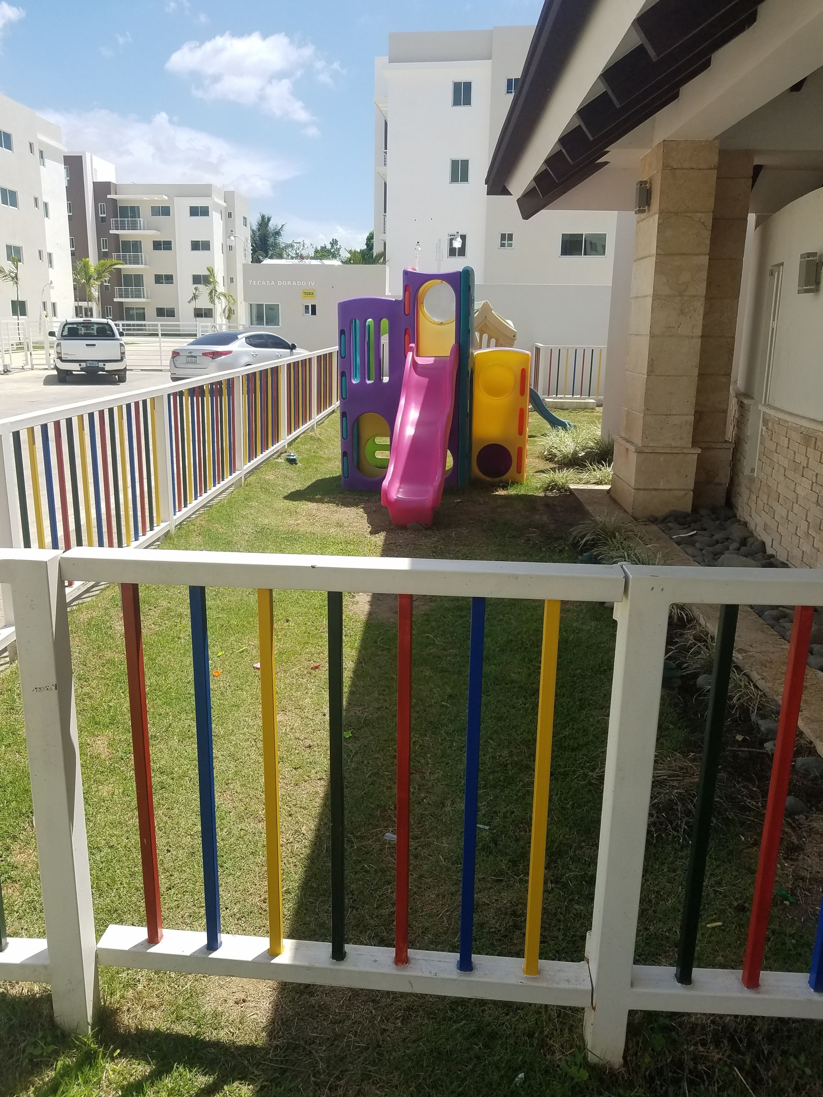 A playground with colorful slides and a fenced area, surrounded by buildings and parked cars.
