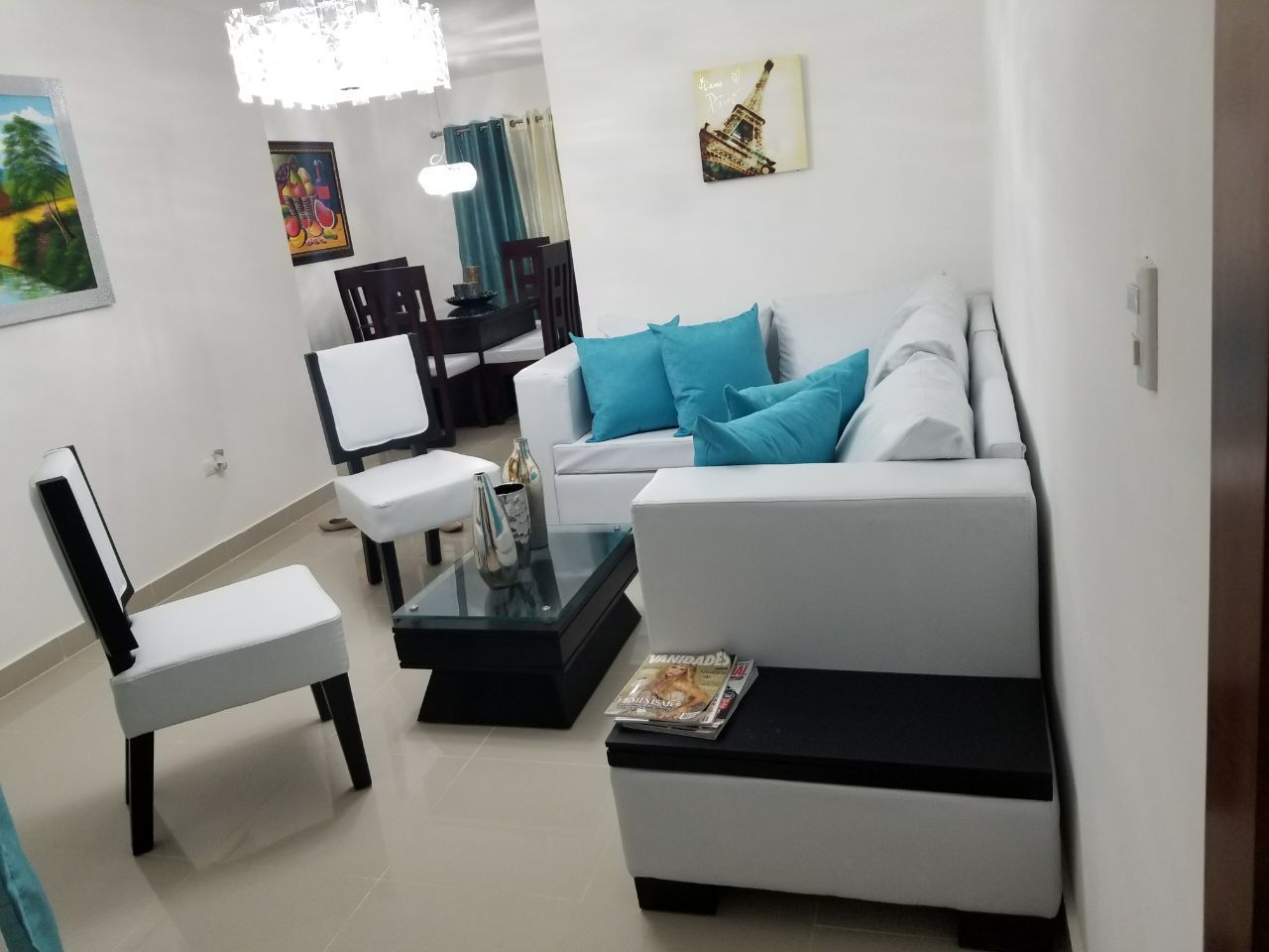 A modern living room with a white sofa, a glass coffee table, and a white armchair. There is a dining area in the background with a table and chairs.