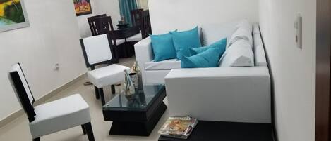 A modern living room with a white sofa, a glass coffee table, and a white armchair. There is a dining area in the background with a table and chairs.