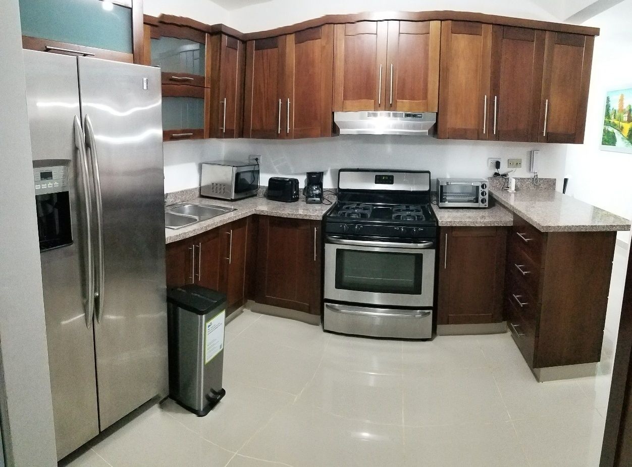 A kitchen with stainless steel appliances, wooden cabinets, a trash bin, and a tiled floor.