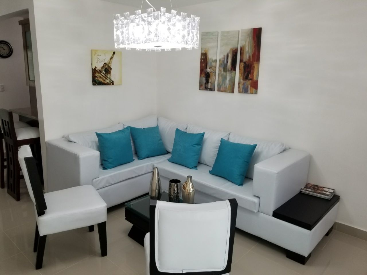 Ein modernes Wohnzimmer mit einem weißen Sofa, Türkis-farbenen Kissen, einem Glastisch und einem weißen Sessel.