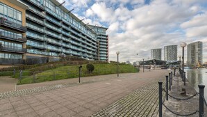 A modern building with multiple floors, a paved walkway, and a body of water in the background. - Premium London Excel Apartment (London)