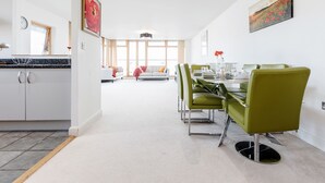 A modern dining area with a glass table and green chairs, a kitchen with white cabinets, and a large window providing natural light. - Premium London Excel Apartment (London)