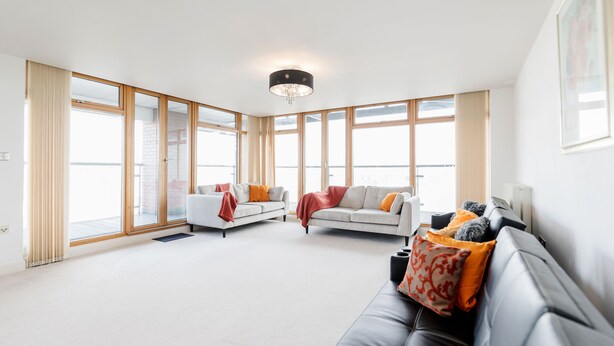 A spacious living room with large windows, a black leather sofa, and a white sofa with orange cushions. - Premium London Excel Apartment (London)