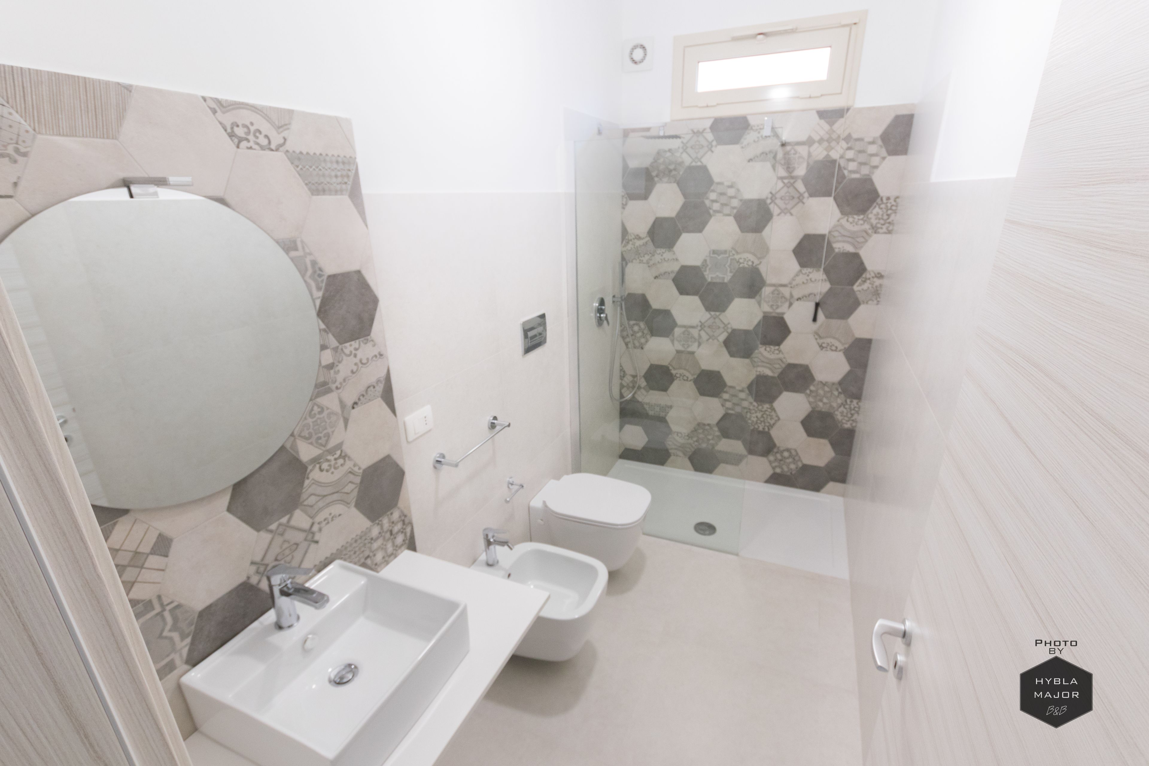 Un bagno moderno con un grande specchio, un lavandino, un bidet e una doccia con parete rivestita di piastrelle decorate.