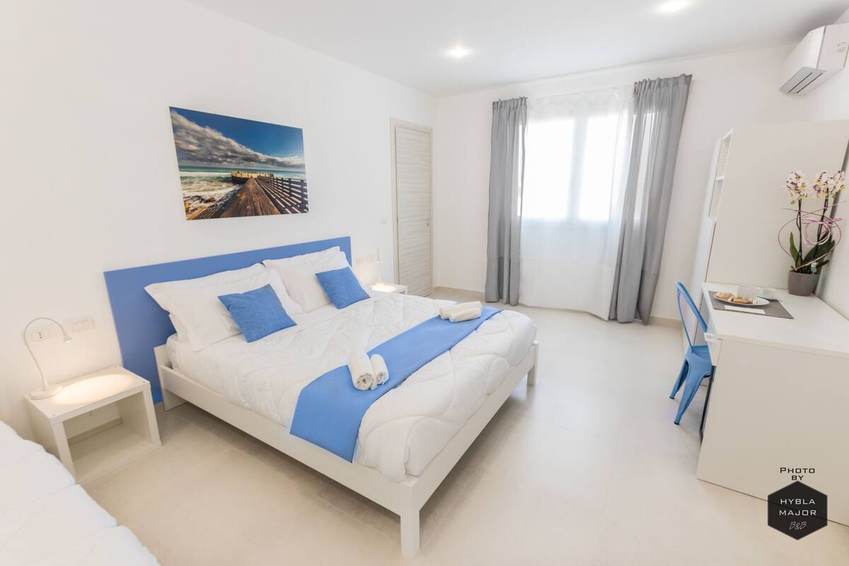 Un dormitorio moderno con cama, escritorio, silla, mesita de noche, un cuadro en la pared y ventana con cortinas.