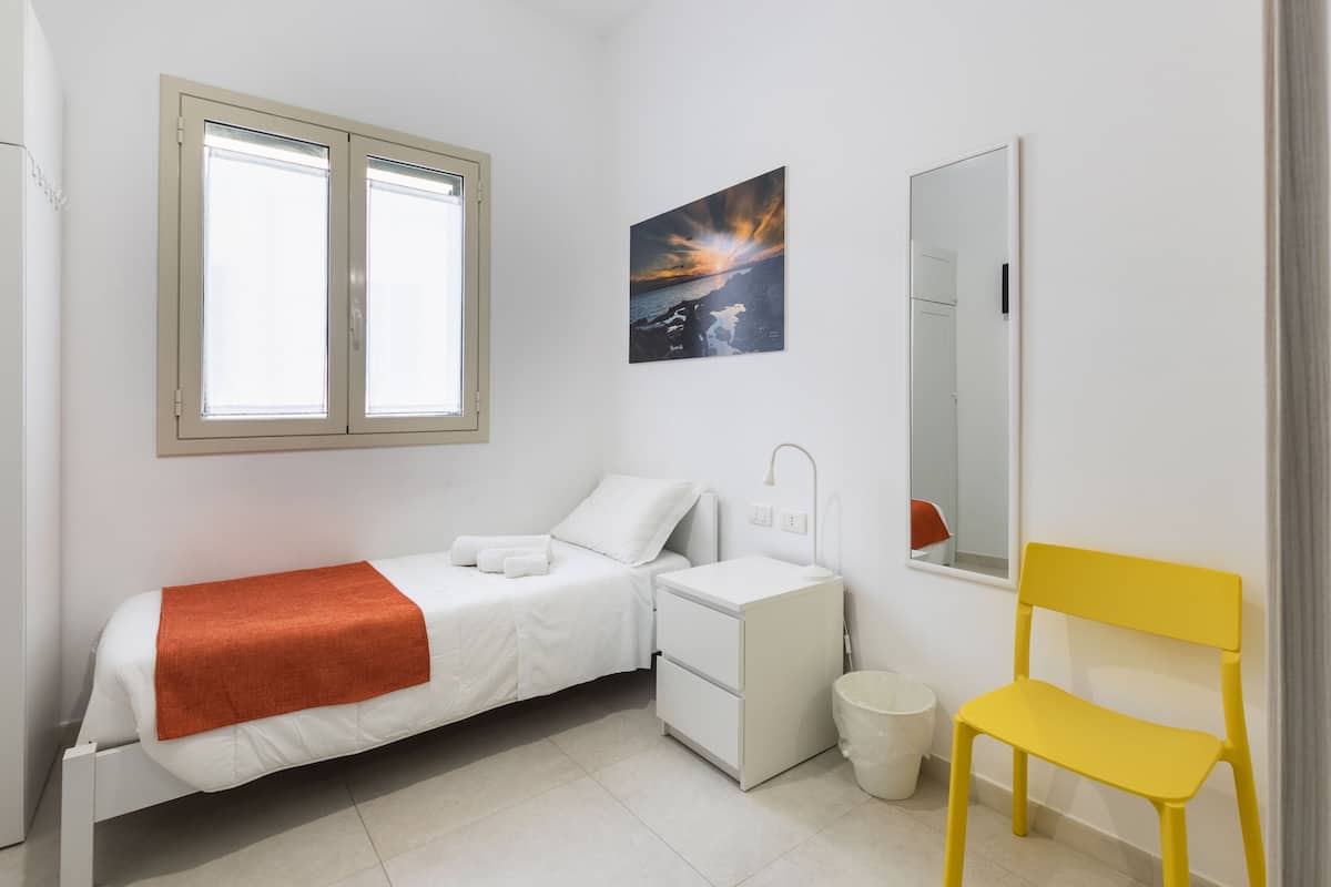 Un dormitorio con cama, mesita de noche, silla, ventana y un cuadro en la pared.