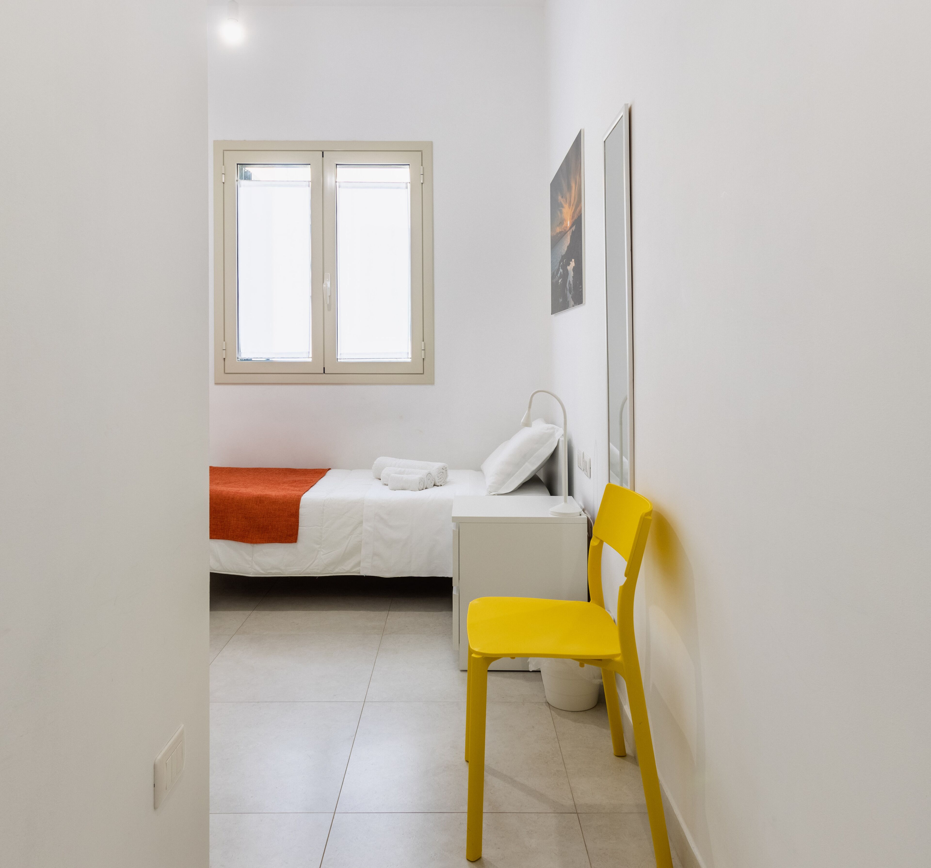 A small, bright room with a bed, a chair, a bedside table, and a window.